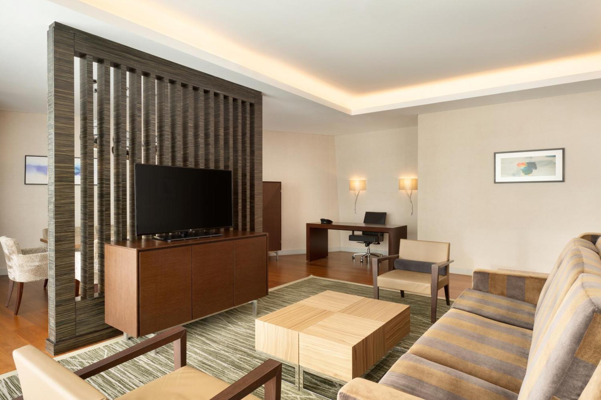 Hilton Riyadh Hotel&Residences Exterior foto The photo shows a modern living space that features a contemporary design. It includes a seating area with a beige sofa and two armchairs arranged around a square wooden coffee table. A wall-mounted flat-screen TV is positioned on a decorative wooden